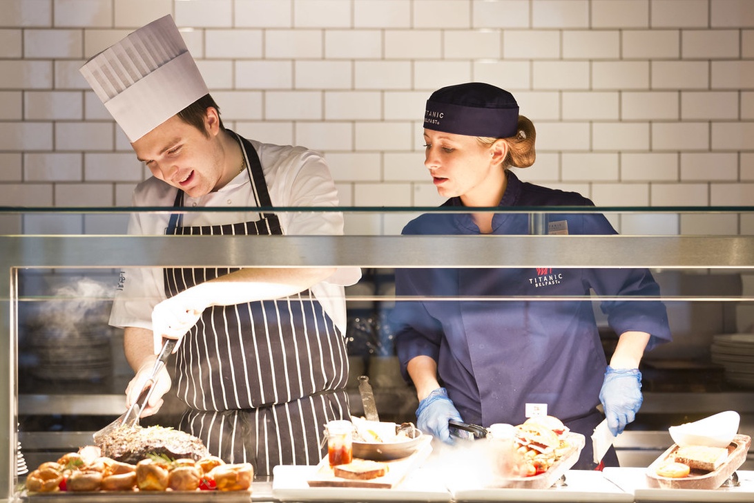 Zwei Restaurantmitarbeiter in einer professionellen Küche bei der Zubereitung von Speisen. Ein Koch in traditioneller Kleidung arbeitet neben einem Mitarbeiter in blauer Uniform mit 'Titanic' Branding.